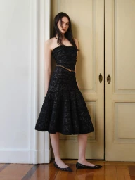 Model standing against pale double doors wearing a strapless black lace midi dress and thin gold belt