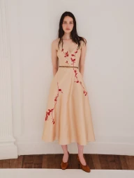 Model in a cream midi dress with red floral embroidery standing against an interior wall