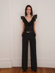 Model Clara Denison wearing a black ruffled lace top and high-waisted wide-leg trousers on wood flooring in front of a white wall.