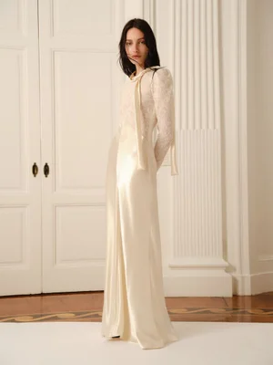 Model wearing a long cream silk dress with lace sleeves standing in front of white paneled doors and a column.