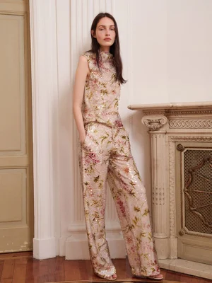 Model Clara Denison wears a sequined floral sleeveless top and wide-leg trousers beside a carved fireplace mantel.