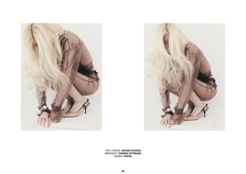 Model with long blonde hair crouching in a sheer brown mesh outfit and kitten heels against a stark white backdrop.