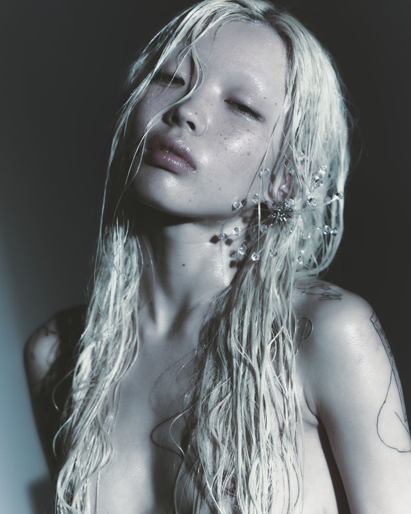 Close-up portrait of an East Asian woman with wet platinum blonde hair, dewy skin, and crystal wire earrings against a dark background.
