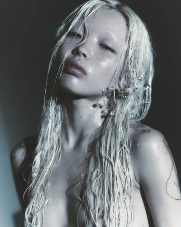 Close-up portrait of an East Asian woman with wet platinum blonde hair, dewy skin, and crystal wire earrings against a dark background.