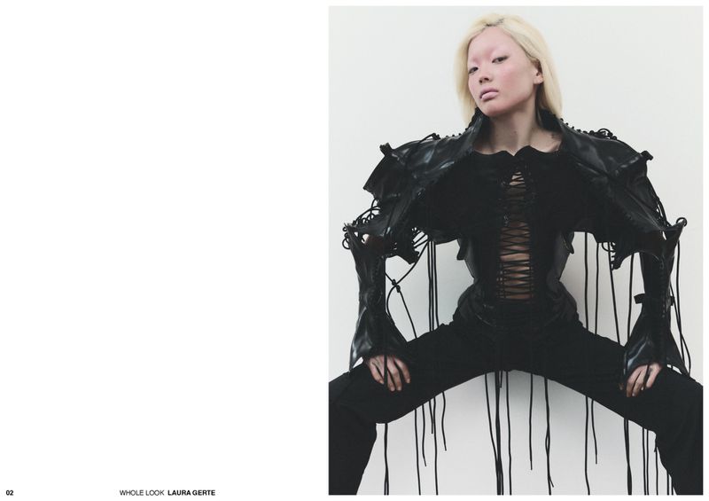 Model with platinum blonde hair wearing a sculptural black corset jacket with trailing cords against a white studio background.