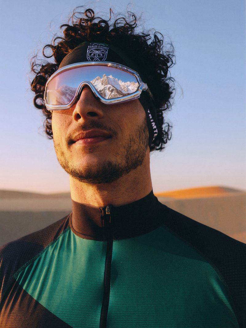 Man in mirrored goggles reflecting snowy peaks wearing a green athletic jersey against a desert sand dune.