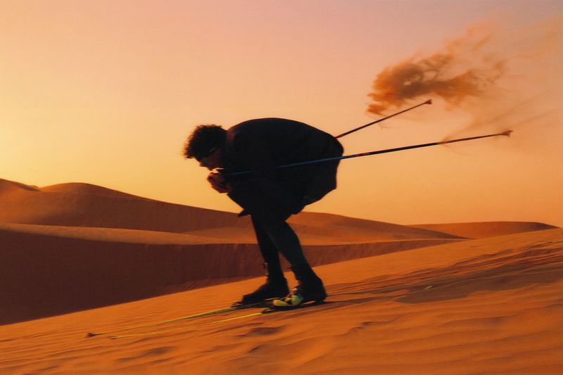 A man in dark apparel crouched in a tuck position skiing down a desert sand dune with ski poles trailing sand under warm sunset light.