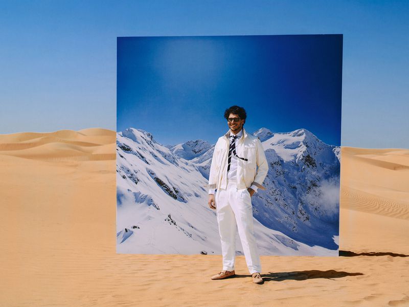 Man in an all-white suit and sunglasses standing in the desert in front of a large backdrop showing snowy mountains.