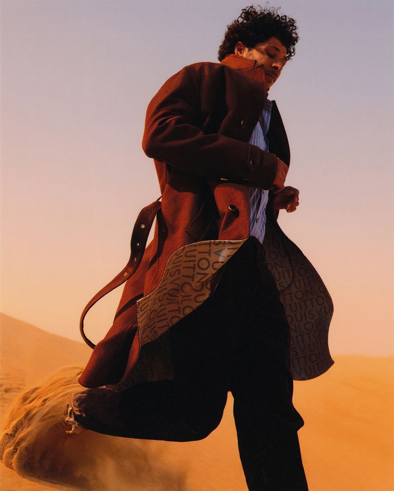 Man running across desert sand dunes wearing a rust trench coat with monogram lining and dark trousers.