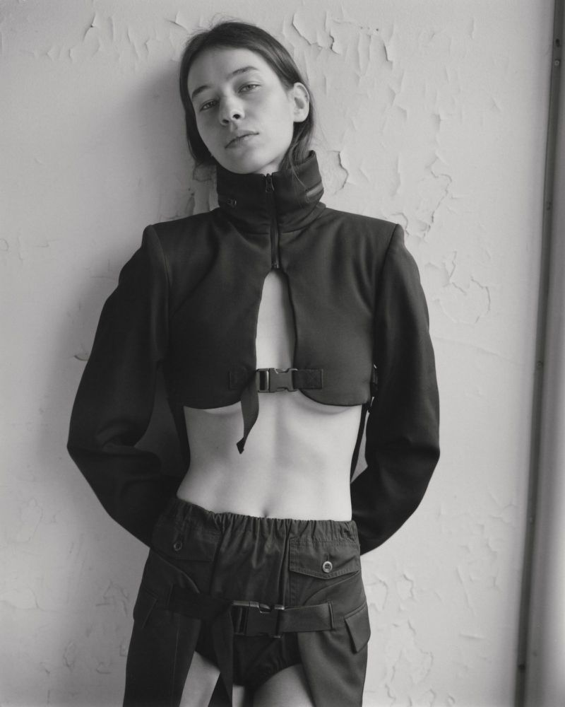 Model Selia Hamdani posing against a textured wall wearing a black cropped high-collar buckle jacket and matching utility bottoms.