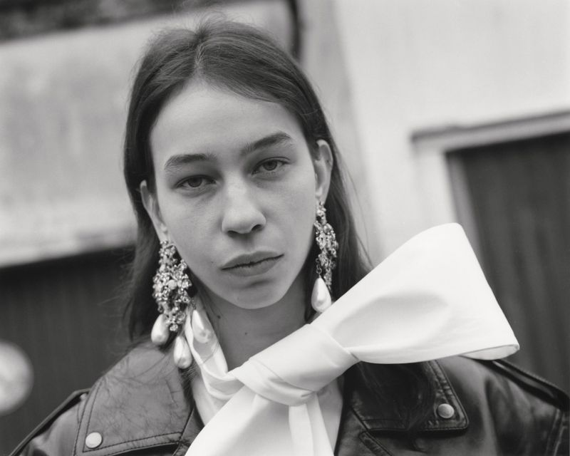Black and white portrait of a woman wearing a leather biker jacket, large white bow scarf, and ornate pearl statement earrings.