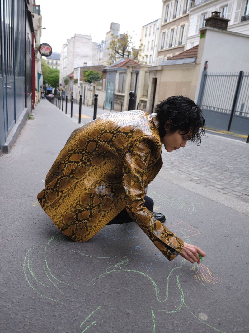 Model Gabrielle Rul wearing a snakeskin print jacket crouching and drawing with chalk on an urban asphalt street.