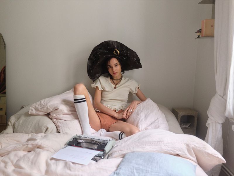 Model Gabrielle Rul wearing an oversized black hat, cream top, and knee-high socks sitting on a bed beside a vintage typewriter.