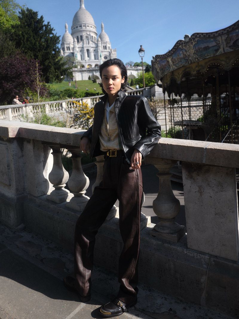 Gabrielle Rul wearing a black leather jacket and brown trousers, leaning on a stone balustrade with the Sacré-Cœur in Paris behind her.