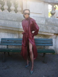 Gabrielle Rul wearing a glossy burgundy belted trench coat and dark sunglasses standing in front of a green park bench.