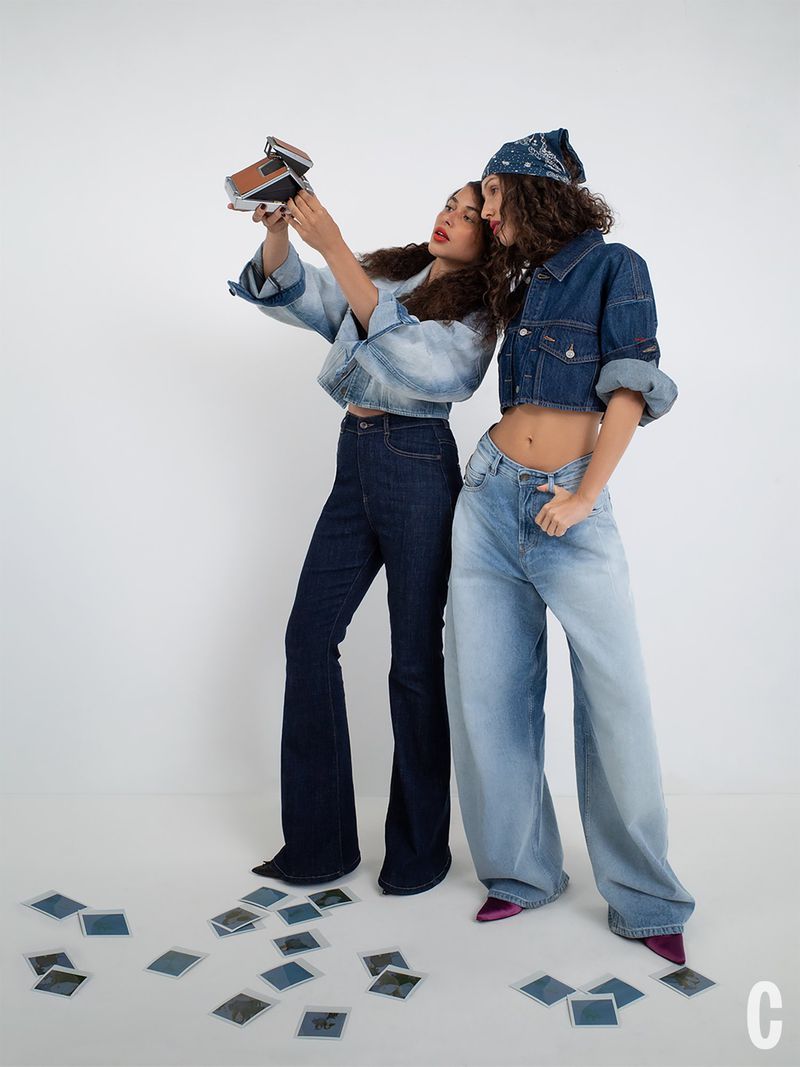 Two models posing in cropped denim jackets and jeans with a vintage instant camera in a white studio setting.
