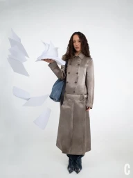 Model Alici Sol stands in a long taupe metallic trench coat with floating sheets of paper against a white studio backdrop.