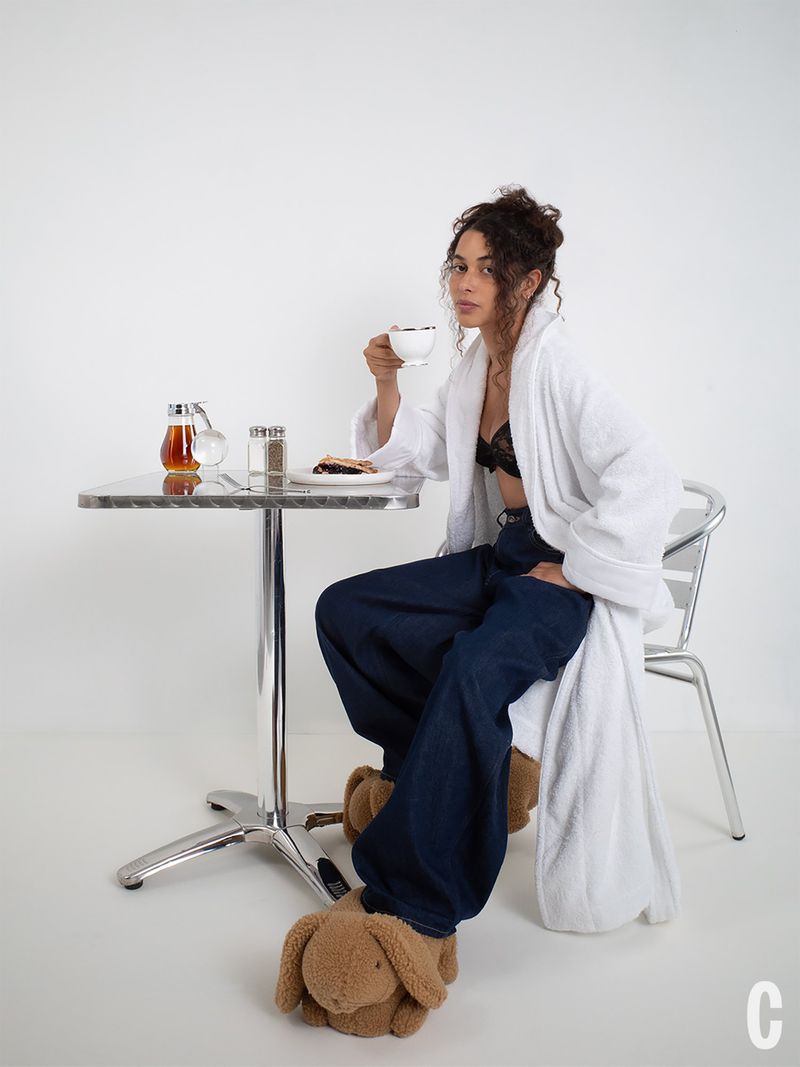 Model Alici Sol in a white bathrobe, wide-leg denim jeans, and plush slippers holding a teacup at a metal cafe table.