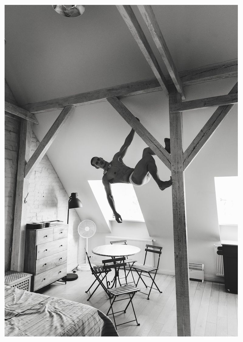 Black and white editorial photo of a man suspended from wooden ceiling beams inside a sunlit attic loft bedroom.