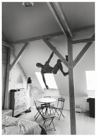 Black and white editorial photo of a man suspended from wooden ceiling beams inside a sunlit attic loft bedroom.