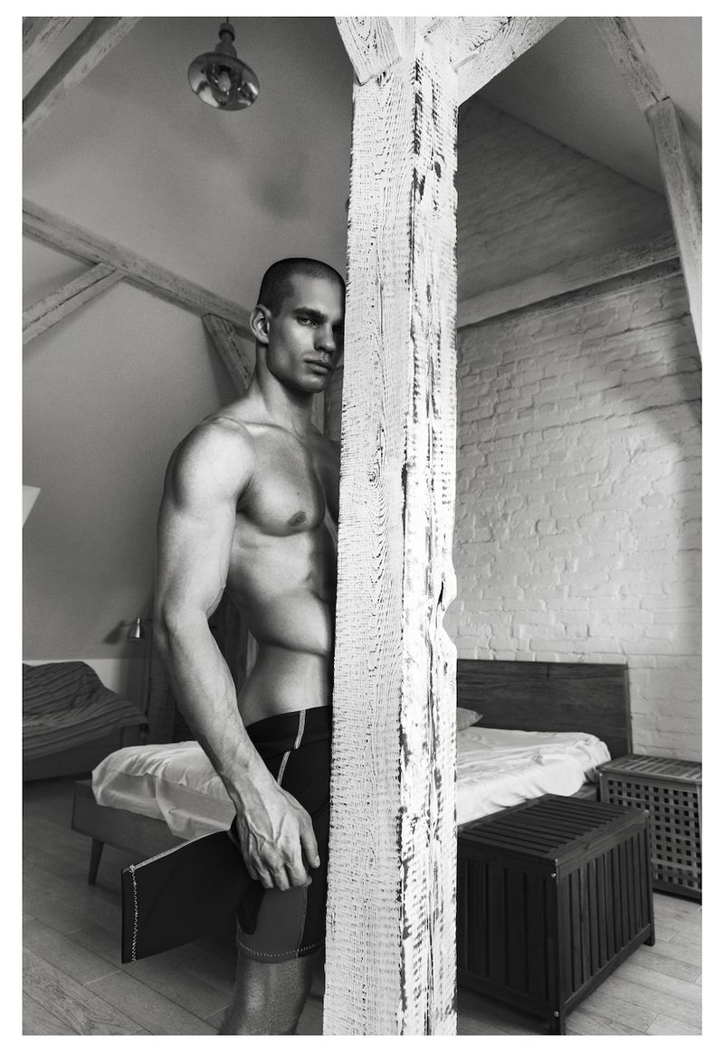 Muscular male model Ivan Zaretski standing shirtless beside a rustic wooden pillar in a loft bedroom setting.