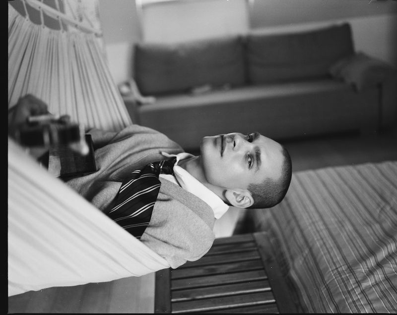 Model Ivan Zaretski lying in an indoor hammock holding a guitar wearing a collared shirt, striped tie, and sweater in black and white.