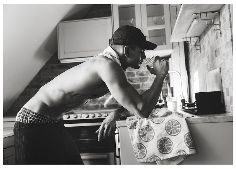 Monochrome shot of a shirtless man wearing a baseball cap and dark trousers leaning against a kitchen counter drinking from a glass.