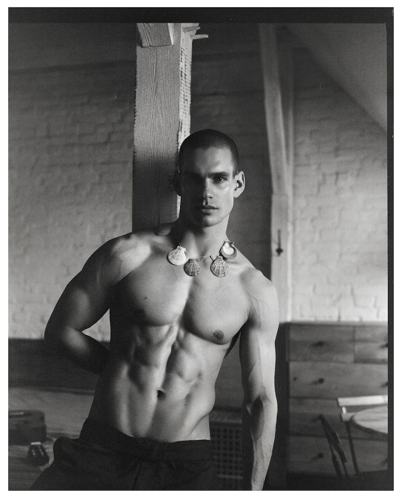 Shirtless muscular man wearing a scallop shell necklace leaning against a wooden pillar in a room in black and white.