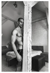 Muscular male model Ivan Zaretski standing shirtless beside a rustic wooden pillar in a loft bedroom setting.