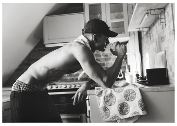 Monochrome shot of a shirtless man wearing a baseball cap and dark trousers leaning against a kitchen counter drinking from a glass.
