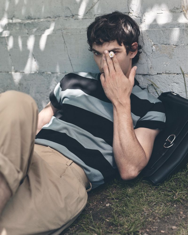 Model Habib Masovic in a striped t-shirt and beige trousers lying on grass against a wall holding a flower to his eye.