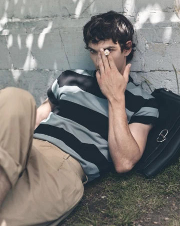 Model Habib Masovic in a striped t-shirt and beige trousers lying on grass against a wall holding a flower to his eye.