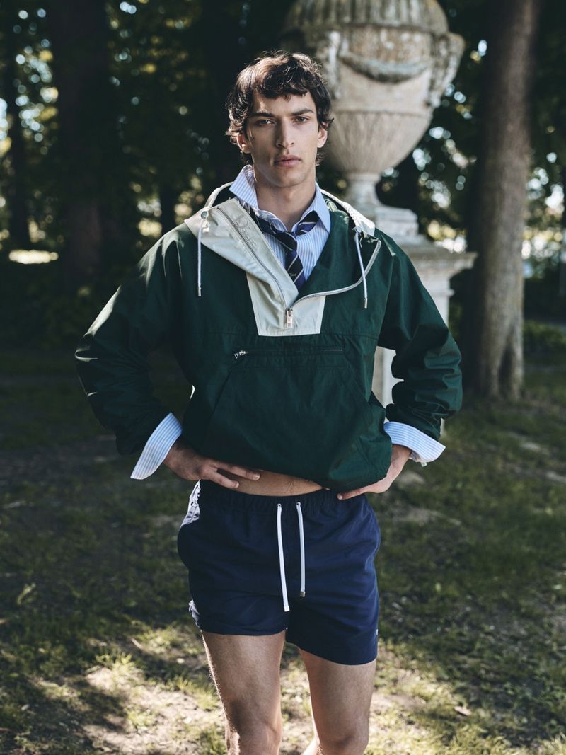 Model Habib Masovic posing outdoors in a green anorak jacket, tie, and navy shorts before an ornate stone garden urn.