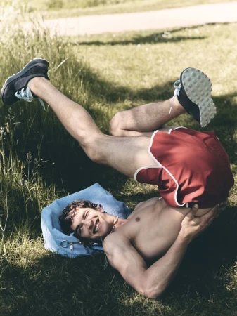 Model Habib Masovic rolls backward onto the grass wearing red retro running shorts and athletic sneakers in a sunlit park.