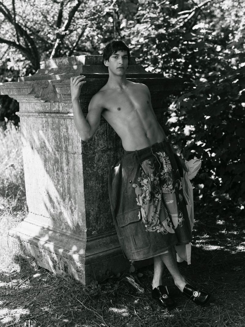 Black and white portrait of a shirtless male model leaning against an aged stone monument in dappled sunlight outdoors.