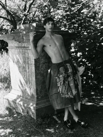 Black and white portrait of a shirtless male model leaning against an aged stone monument in dappled sunlight outdoors.