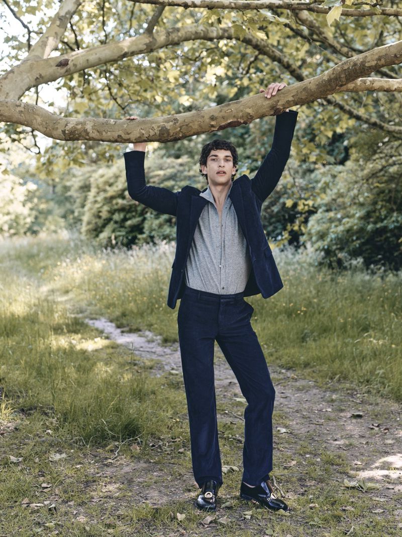 Model Habib Masovic holding a low tree branch wearing a navy blue tailored suit and grey shirt in an outdoor park setting.