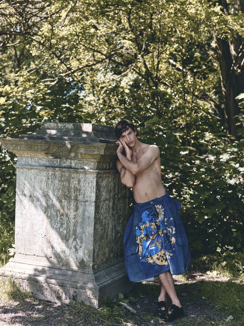 Shirtless male model in printed blue denim shorts leaning against an aged stone pedestal in a wooded park.