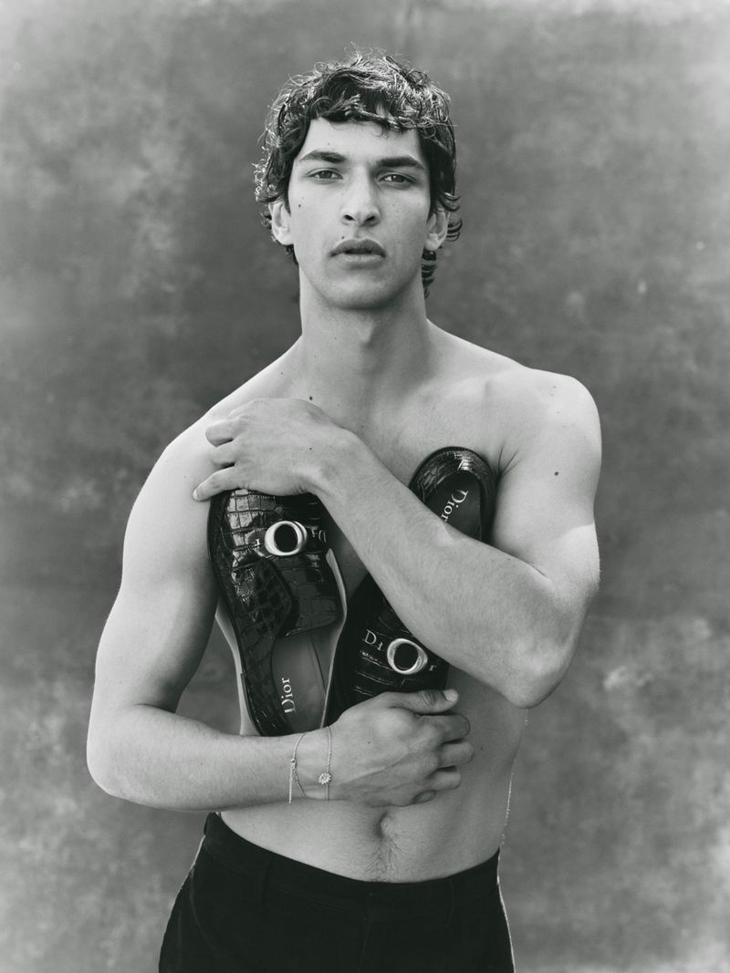 Shirtless male model Habib Masovic holds black crocodile-embossed Dior loafers across his chest against a textured backdrop.