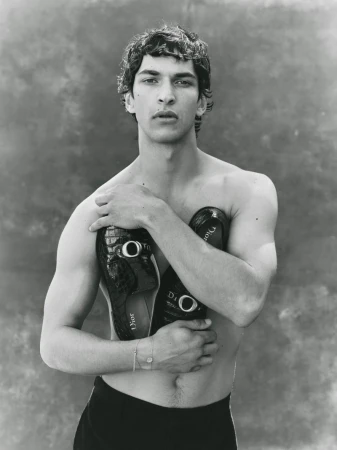 Shirtless male model Habib Masovic holds black crocodile-embossed Dior loafers across his chest against a textured backdrop.