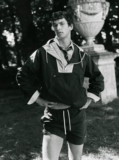 Model Habib Masovic wearing a hooded anorak, collared shirt with necktie, and shorts outdoors beside a classical stone urn.