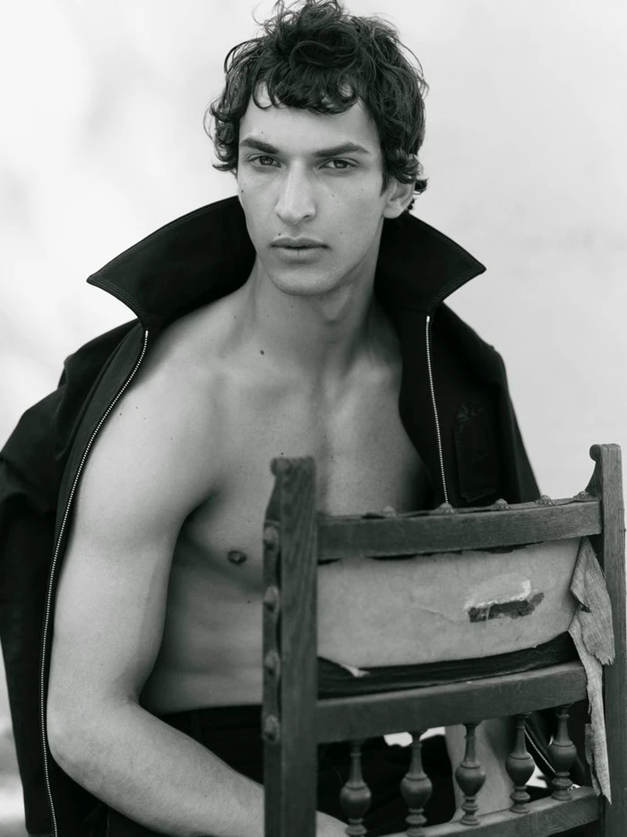 Black and white portrait of Habib Masovic shirtless wearing an open black jacket, seated backwards on a vintage wooden chair.