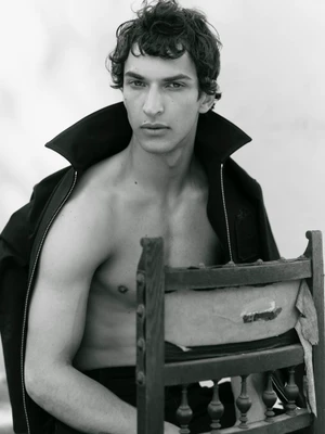 Black and white portrait of Habib Masovic shirtless wearing an open black jacket, seated backwards on a vintage wooden chair.