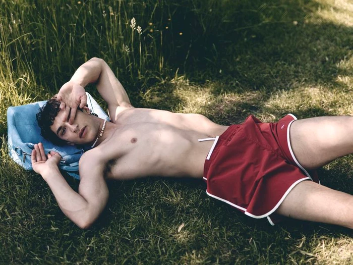 Model Habib Masovic shirtless in red swim shorts lying on green grass with a light blue bag behind his head.