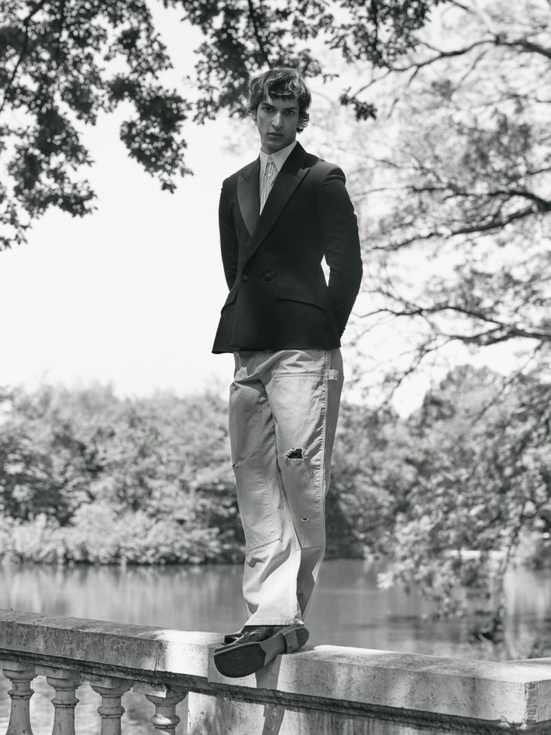 Model Habib Masovic standing on a stone balustrade wearing a black blazer and distressed trousers beside a lake in black and white.