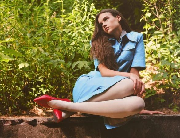 Young woman in a blue dress and red shoes seated outdoors surrounded by green foliage