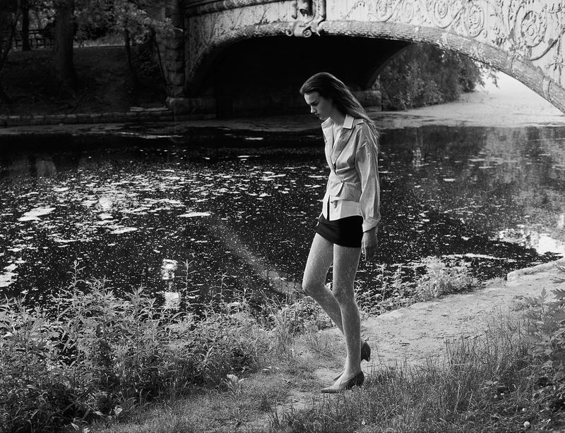 Woman walking along a dirt path beside water near an ornate arched bridge wearing a blouse, black miniskirt, and tights in black and white.