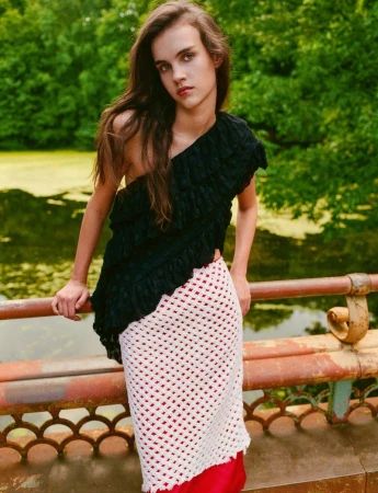 Model leaning on an ornate bridge railing wearing a black one-shoulder ruffled top and a white open-knit crochet skirt.