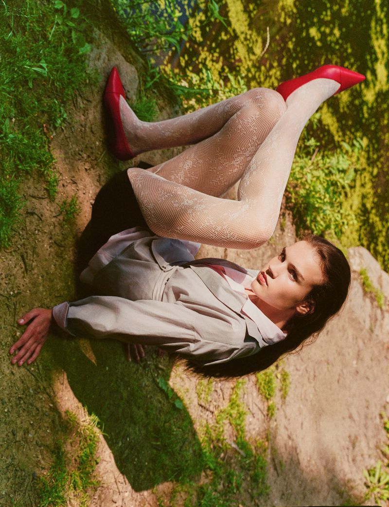 Model in a light grey blazer, white patterned lace tights, and red pointed-toe pumps reclining outdoors in dappled sunlight.