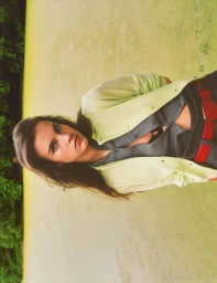 Model wearing a pale lime green cardigan, grey shirt, and bold red belt posing outdoors against an algae-covered pond.